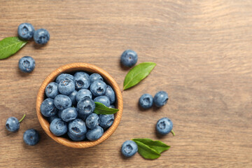 Bowl of tasty fresh blueberries with green leaves on wooden table, flat lay. Space for text