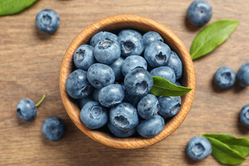 Bowl of tasty fresh blueberries with green leaves on wooden table, flat lay