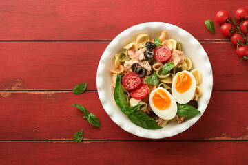Italian pasta salad. Orecchiette pasta with tuna, tomato cherry, olive, basil and parmesan cheese in plate on red old rustic table background. Traditional Italian cuisine. Flat lay. Copy space.