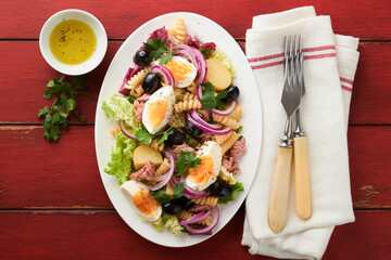 Tuna salad with pasta, eggs, potatoes, olives, red onions and sauce in white plate on old red rustic table background. Nicoise salad. French cuisine. Top view. Flat lay.