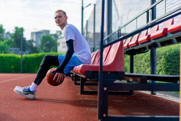 A tall guy basketball player sits in the bleachers of an outdoor basketball court and demonstrates dribbling a basketball before practice starts 