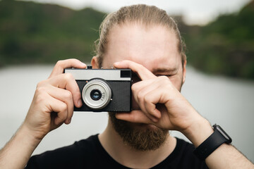 A young male photographer takes a photo on an old film camera on a blurred background of a river in a mountainous area, the concept of tourism.