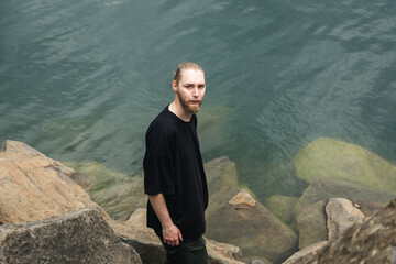 A young man with a beard and a bun in a gray T-shirt stands among the stones near the lake or the...