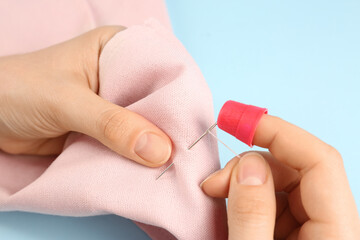 Woman sewing on pink fabric with thimble and needle against light blue background, closeup