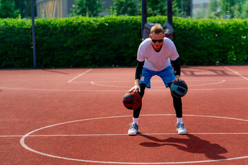 Obraz premium A tall guy basketball player with two balls shows off his dribbling skills while practicing on a basketball court in the park 