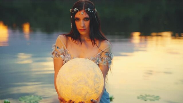 Fantasy woman holding glowing moon planet in hands. Girl fairy tale elf beauty face look at camera, river nymph wet creative dress, stands in water dark lake summer nature. Lady queen night. art 4k