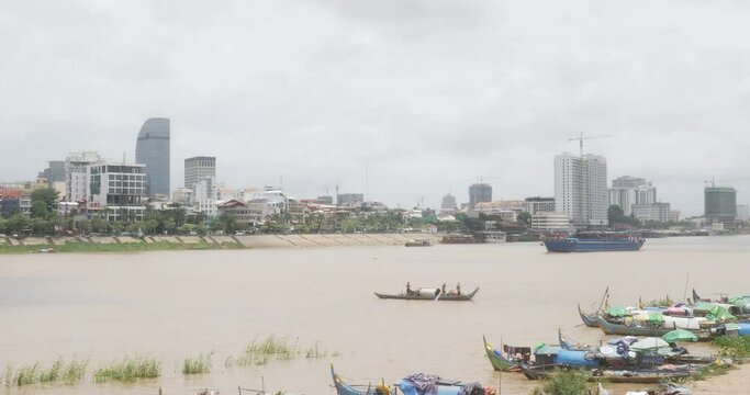 A row of traditional fishing boats moored along the river bank in the foreground. Fishers in a small boat pulling up a net on the Mekong river. Empty barge sailing as backdrop