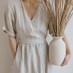 Young beautiful woman in neutral beige creamy linen dress holding sandy earthenware with dried...