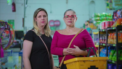 Portrait of two serious female customers standing inside grocery store holding basket in hand. Middle-aged women posing for camera at supermarket with worried stern expression