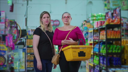 Portrait of two serious female customers standing inside grocery store holding basket in hand. Middle-aged women posing for camera at supermarket with worried stern expression