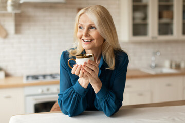 Happy morning. Happy mature woman drinking coffee in kitchen, standing with cup in hands, looking aside and smiling