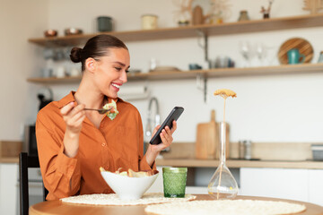 Happy european woman eating tasty salad and using cellphone in kitchen while having dinner, sitting...