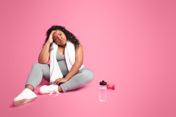 Tired black obese lady in sportswear, towel on shoulders, sitting on floor with dumbbells, bottle of water, free space