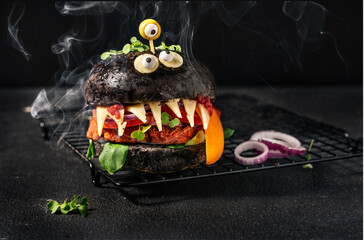 Monster burger with black bun, vegan patty, cheese, olive eyes and smoke on the black background....