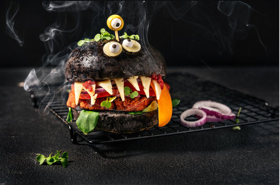 Monster burger with black bun, vegan patty, cheese, olive eyes and smoke on the black background. Halloween food
