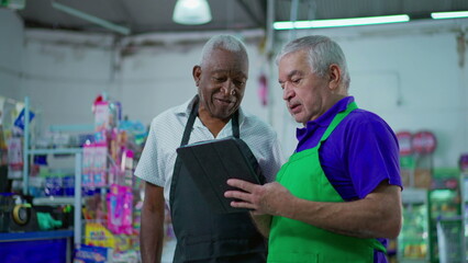 Obraz premium Two happy diverse senior staff of frocery store checking inventory operations with tablet device, older employees of supermarket laughing and smiling at workplace