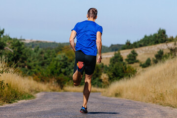 rear view male runner in blue shirt and black tights running road in field of dry grass