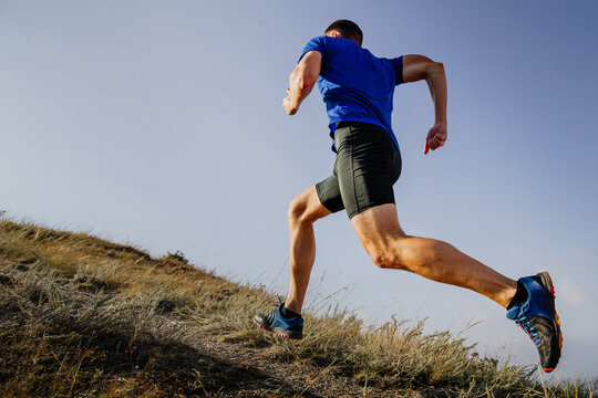 athlete runner running mountain uphill in blue shirt and black tights, background of sky