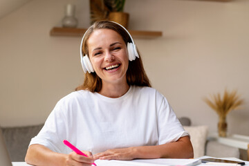 Front view portrait of young laughing female student in headphones wearing white tee shirt doing homework. Study, learning, education.