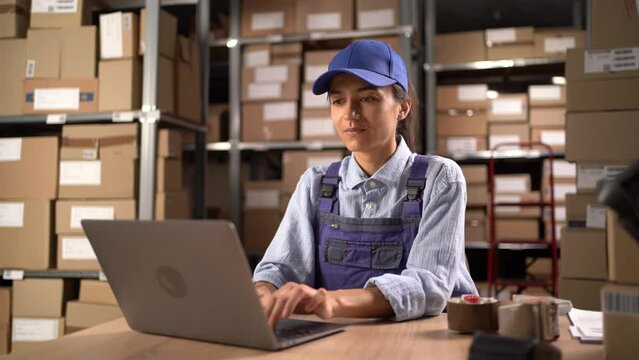 Female worker in overalls sitting and working using laptop computer in storage warehouse. People, warehouse and industry