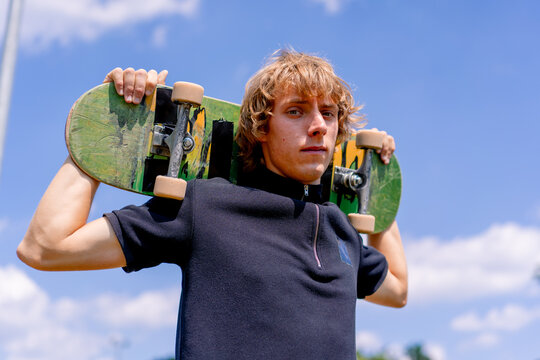 Portrait of a young serious guy skater with long hair holding a skateboard on his shoulders on the background of skatepark and sky
