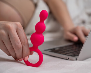 Woman holding pink anal beads next to laptop while lying on bed. 