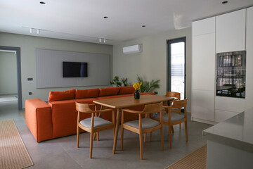 Open plan living room with orange textile couch and white cupboard kitchen with built-in...