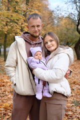 Young parents, mother and father, walk with a newborn baby in the autumn park. Parents smiling and looking at the camera