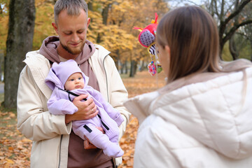 Young mother and father are walking with a newborn baby in the autumn park. The father holds the child in his arms, the mother shows her a toy