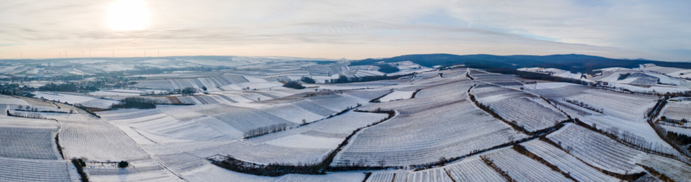 Aerial panoramic drone view in winter of vineyards covered in snow as the sun sets with wind turbines in the far distance on a cloudy day, Poysdorf, Lower Austria, Austria.