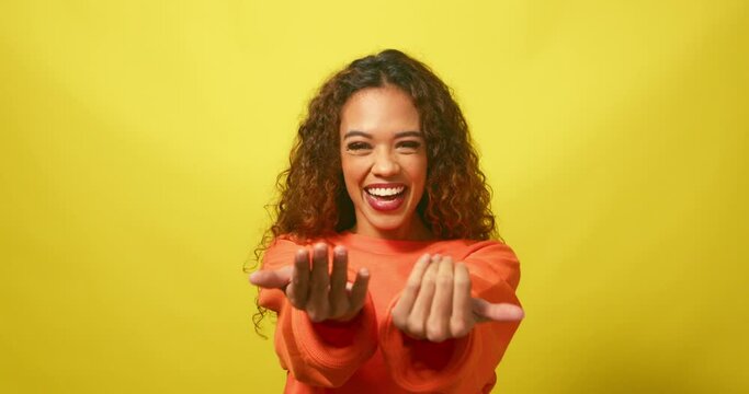 Flirty young woman beckons with hands 'come here', yellow studio background