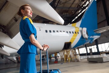 Pretty air stewardesses in bright blue uniform smiling away while standing with suitcase in front of passenger aircraft. Occupation concept