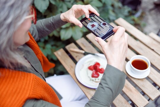 Senior woman guest takes picture of beautiful strawberry dessert with smartphone sitting at table on outdoors cafe terrace on autumn day