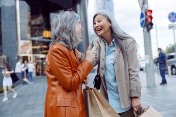 Cheerful mature women in stylish outfit with shopping bags talk on modern city street on autumn day. Friends spend time together