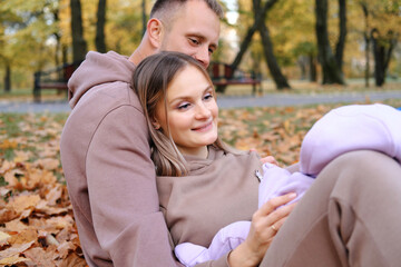 Naklejka premium Young parents sit on a plaid with a newborn baby in the autumn park. The child lies on the lap of the mother, the parents play with the child