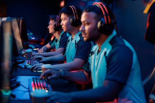 Group of focused esports players sitting together in front of computer monitors and playing video game in internet cafe