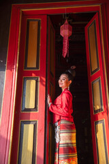 Close up young Asian woman with Thai Lanna traditional dress stand at door with red color and old style of ancient house also look to left side with smiling.
