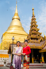 Vertical image of two women stand in front of golden pagoda and ancient building of the temple in Thailand and they also smile to camera.