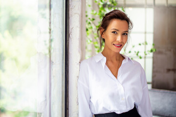 Portrait of an attractive mid aged woman wearing white shirt and black pants while standing by the window
