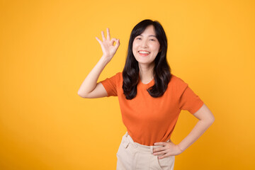Positive Asian woman in her 30s dons orange shirt, showing okay sign on yellow backdrop. Hands...