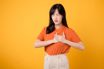 Caring young Asian woman in her 30s, wearing an orange shirt, holds hands on chest on yellow background. heart attack disease, chest pain health care concept.