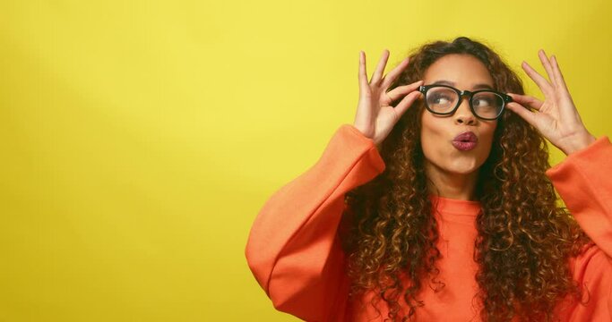Beautiful multi-ethnic woman pulls silly face trying eyeglasses, yellow studio