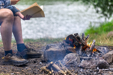Close-up of bonfire with man reading a book in the background in nature.