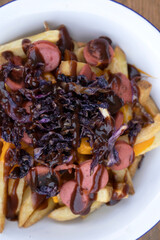 Top view of fried potatoes with sausages, red cabbage, cheddar cheese and barbecue sauce, in a white bowl with a wooden background.	
