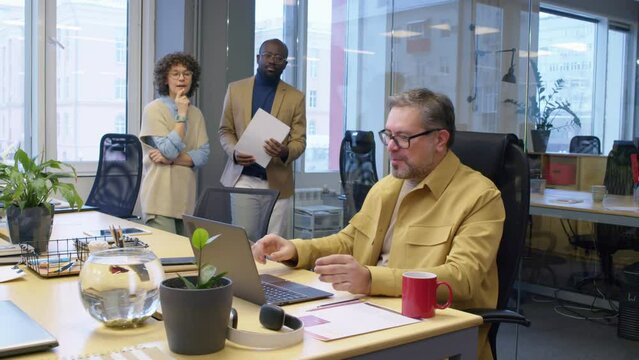 Medium full shot of happy male Caucasian manager sitting at desk in office showing results of project to supportive diverse coworkers