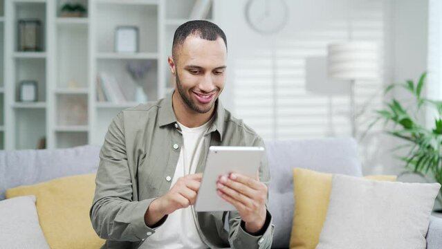 Young adult man using digital tablet while sitting at home office in living room Happy Male guy freelancer businessman or entrepreneur typing browsing application in smartphone device working remotely