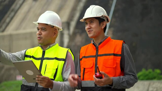 Confident asian two maintenance engineers man inspection with in tablet at construction site dam with hydroelectric power plant and irrigation. Team engineer man working at project big building.and ir
