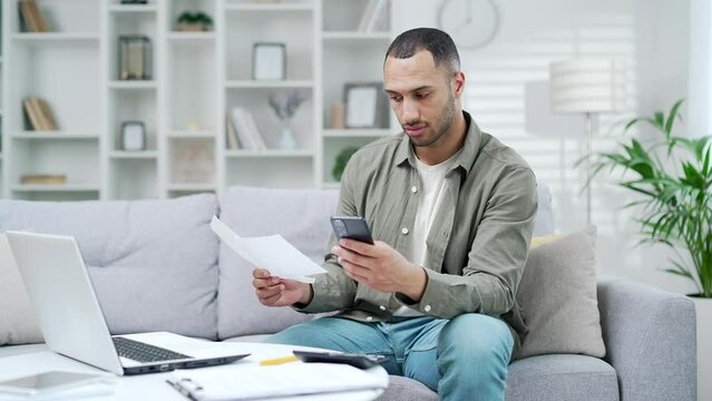 Happy man uses mobile app for spending bills using smartphone on phone while sitting at home in room. A male calculates household finances and enters data into a chat bot easy and simple to pay 