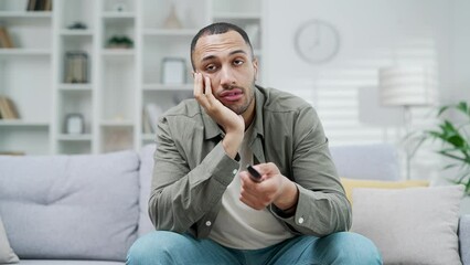 Bored male guy at home in front of TV Exhausted sad young adult man holds a remote control in his hands and switches channels after hard day need some relax indoor house. Boring TV shows films movie