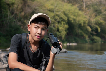 Young asian boy holding binoculars, sitting on rock beside the river in local national park to watch fish in river and birds on the trees and sky.
